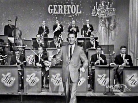 tv show host with people playing music behind him